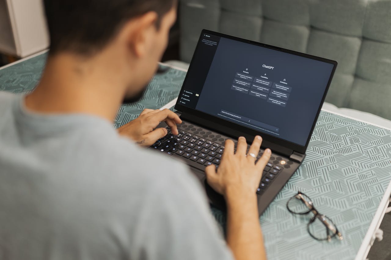 gallery-05 Man using laptop with AI interface, typing with focused attention. Indoors with eyewear beside.